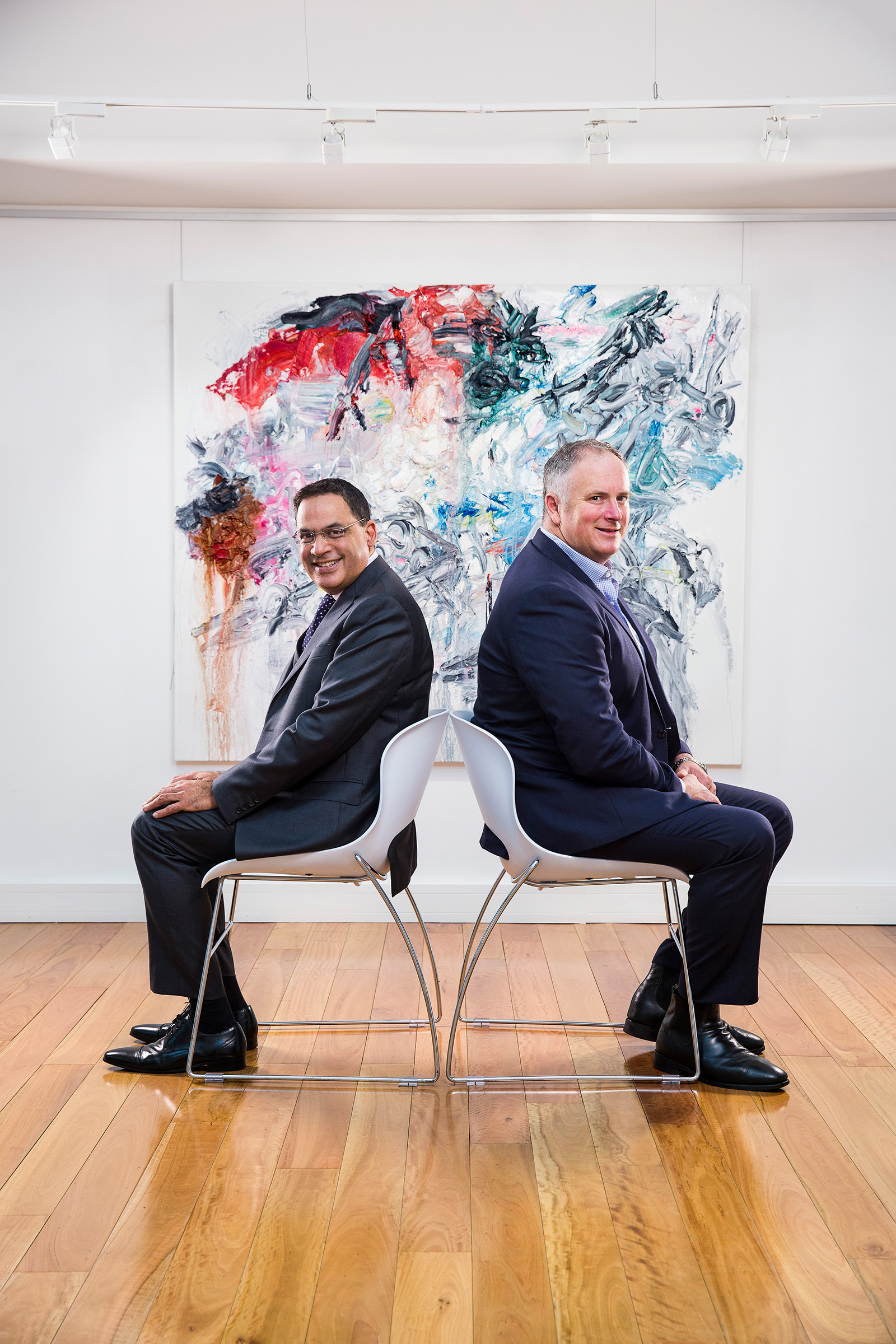 Corporate editorial photography of 2 seated men on white chairs in art gallery with painting in background